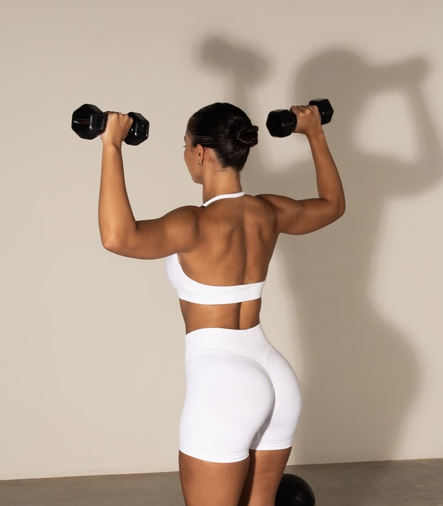 Woman seen from behind in white workout clothes lifting two dumbbells
